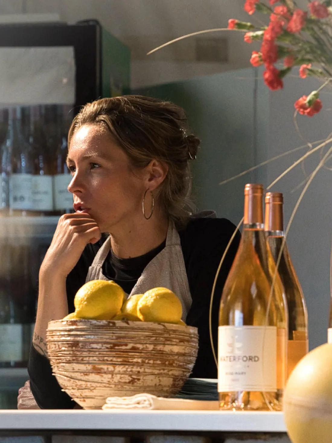 Ragnhild ved disken med en skål citroner og vinflaskerne på række.